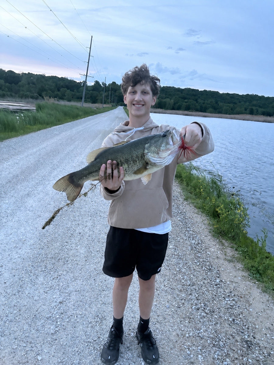 Smiling angler holding a largemouth bass on a gravel road beside a lake